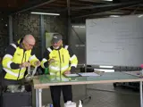 2 Mannen Staan Bij De Innamebalie Kringloopspullen. Ontwikkelcoach Bob Geeft Een Uitleg.